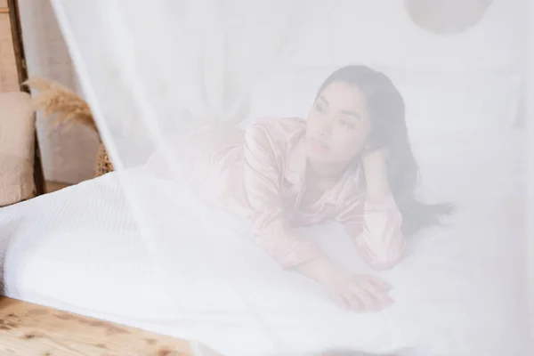 Jovem mulher asiática em pijama de seda deitado fora cortina em linho branco com mão perto da cabeça no quarto — Fotografia de Stock
