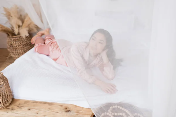 Jeune femme asiatique en pyjama de soie couché dehors rideau sur linge blanc dans la chambre — Photo de stock