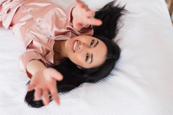 Vista superior de la joven mujer asiática acostada en ropa blanca con las manos extendidas y mirando a la cámara en el dormitorio - foto de stock