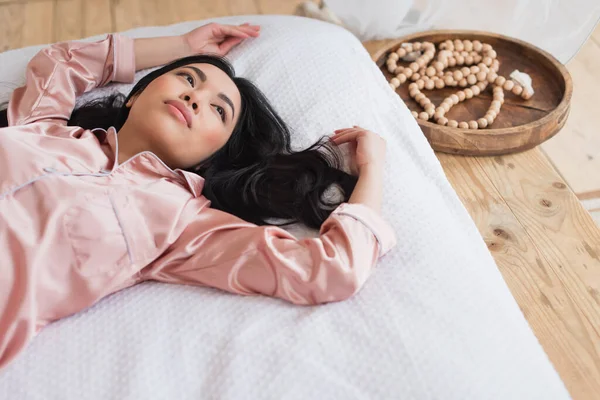 Alto ángulo vista de soñadora joven asiática mujer acostada en blanco lino con las manos extendidas en dormitorio - foto de stock