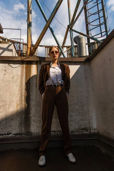 Stilvolles junges Model in braunem Anzug posiert mit Händen in Taschen auf dem Dach — Stockfoto