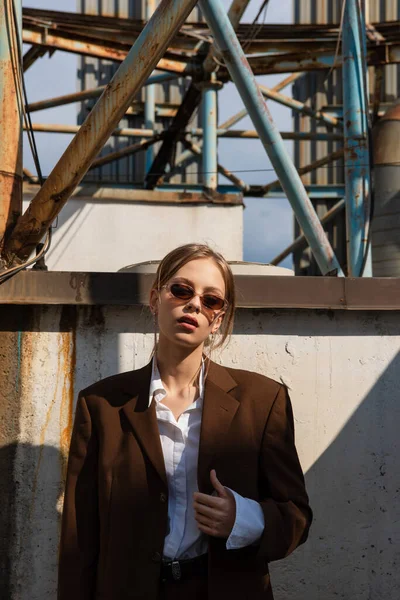 Blonde jeune modèle en costume marron et lunettes de soleil posant à l'extérieur — Photo de stock