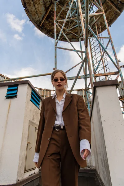 Low angle view of blonde model in sunglasses and trendy brown suit walking on rooftop — Stock Photo