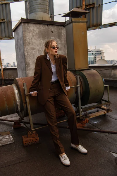 Model in voller Länge mit Sonnenbrille und trendigem braunen Anzug posiert auf dem Dach — Stockfoto