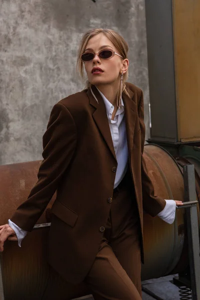 Jeune modèle blonde en lunettes de soleil et costume marron tendance posant sur le toit — Photo de stock