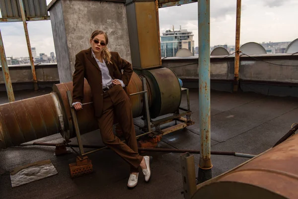 Full length of young model in sunglasses and trendy suit posing with hand on hip and crossed legs on rooftop — Stock Photo