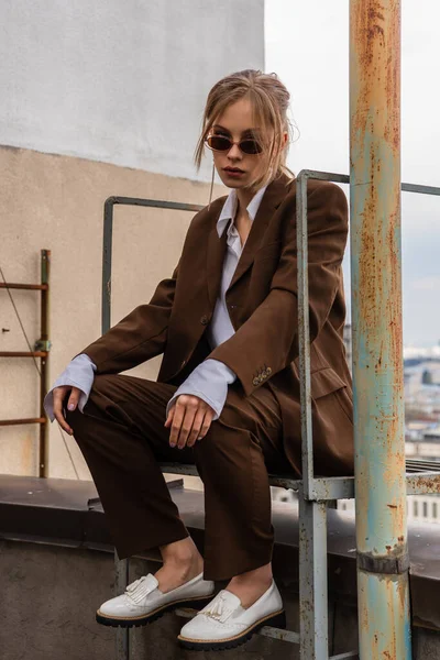 Полная длина молодой модели в стильных солнцезащитных очках и модный костюм, сидящий на крыше — стоковое фото