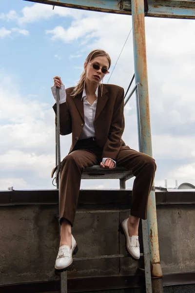 Full length of young model in stylish sunglasses and trendy suit sitting on metallic stairs on rooftop — Stock Photo