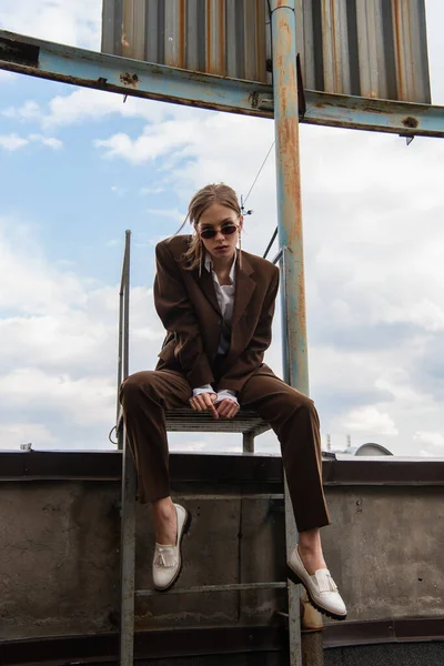 Full length of young woman in sunglasses and trendy suit sitting on metallic rusty stairs on rooftop — Stock Photo