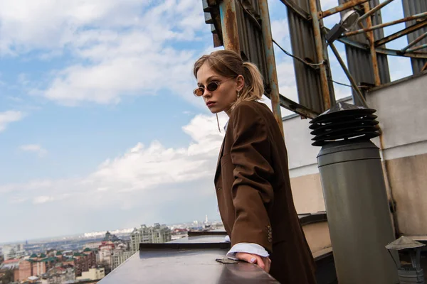 Pretty model in stylish sunglasses posing on rooftop with cityscape on blurred background — Stock Photo