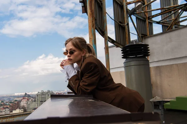 Nachdenkliches Model in stylischer Sonnenbrille posiert auf dem Dach mit Stadtbild vor verschwommenem Hintergrund — Stockfoto