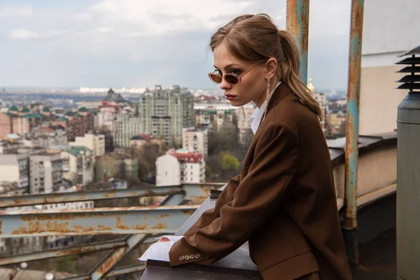 Modelo reflexivo en gafas de sol elegantes posando en la azotea con paisaje urbano sobre fondo borroso - foto de stock