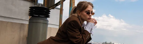 Modelo reflexivo en gafas de sol elegantes posando en la azotea con paisaje urbano sobre fondo borroso, bandera - foto de stock