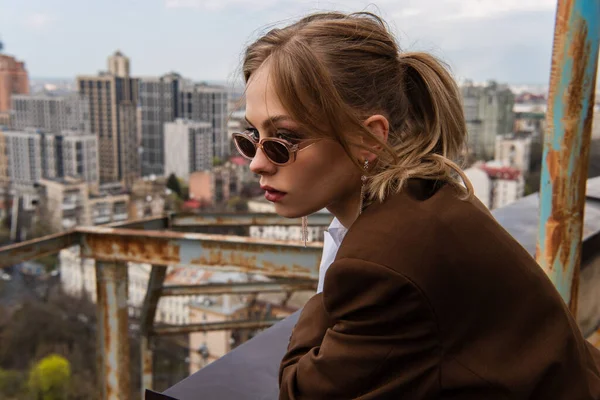Thoughtful woman in stylish sunglasses posing on rooftop with blurred cityscape on background — Stock Photo