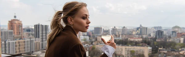 Jovem segurando copo de vinho branco e posando com paisagem urbana no fundo borrado, banner — Fotografia de Stock