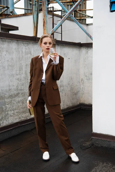 Comprimento total da jovem mulher em elegante terno segurando garrafa e beber vinho no telhado — Fotografia de Stock