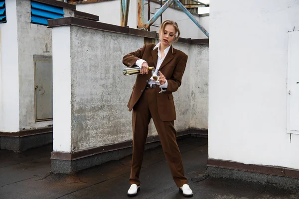 Full length of young woman in stylish suit holding bottle and pouring wine in glass on rooftop — Stock Photo