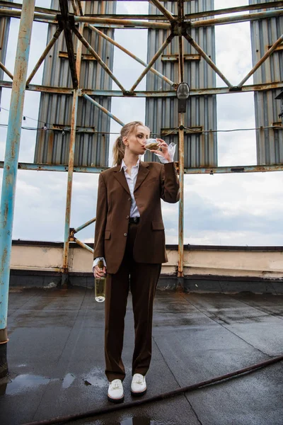 Full length of young woman in stylish suit holding bottle while drinking white wine on rooftop — Stock Photo