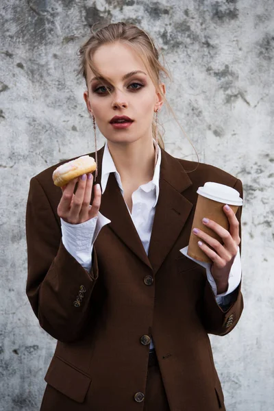 Молодая женщина в стильном наряде держит бумажный стаканчик и пончик к бетонной стене — стоковое фото