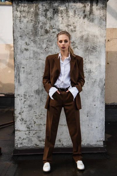 In voller Länge junge Frau in stilvollem Outfit posiert mit Händen in Taschen nahe Betonmauer — Stockfoto