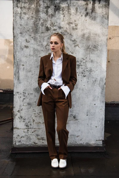 Full length of young woman in stylish suit posing with hands in pockets near concrete wall — Stock Photo