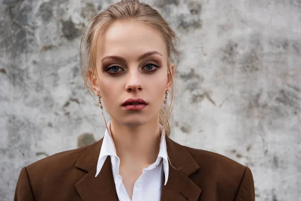 Jovem mulher em brincos elegantes e blazer posando perto da parede de concreto — Fotografia de Stock