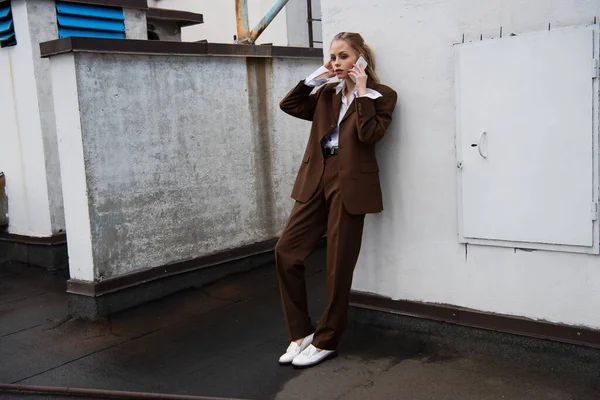 Comprimento total da jovem mulher em roupa elegante falando no celular perto da parede no telhado — Fotografia de Stock