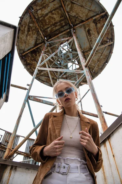 Low angle view of trendy model in blue sunglasses posing near construction on rooftop — Stock Photo