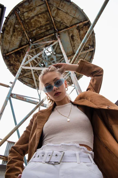 Low angle view of young model in blue sunglasses posing near construction on rooftop — Stock Photo