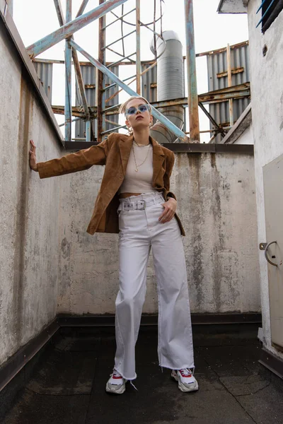 Full length of young woman in stylish outfit with suede blazer and sunglasses posing on rooftop — Stock Photo