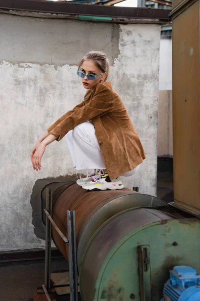 Full length of young woman in stylish outfit with suede blazer and blue sunglasses posing on rusty construction — Stock Photo