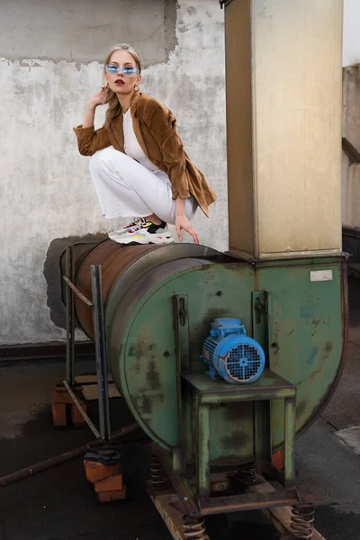 Full length of young model in stylish outfit and blue sunglasses posing on rusty construction — Stock Photo