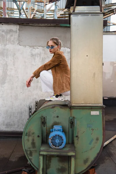 Comprimento total da jovem mulher em roupa elegante e óculos de sol azuis posando na construção enferrujada — Fotografia de Stock