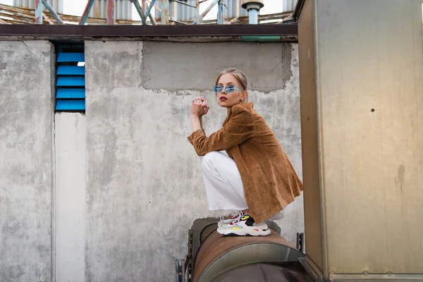 Full length of young woman in trendy outfit and blue sunglasses posing on rusty construction — Stock Photo