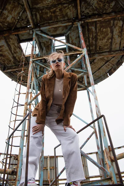 Baixo ângulo de visão do modelo em óculos de sol azuis, calças brancas e jaqueta de camurça posando perto de construção no telhado — Fotografia de Stock