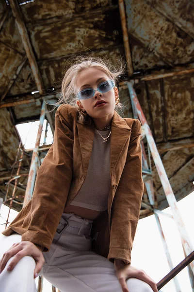 Low angle view of pretty young model in blue sunglasses, white pants and suede jacket looking at camera near construction on rooftop — Stock Photo