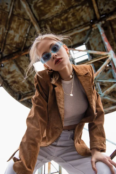 Vue à angle bas de joli jeune modèle en lunettes de soleil bleues regardant la caméra près de la construction âgée sur le toit — Photo de stock