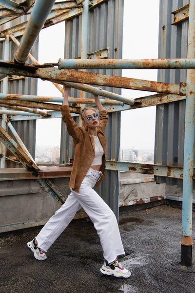 Comprimento total do modelo jovem na moda em óculos de sol azuis posando perto de construção enferrujada no telhado — Fotografia de Stock