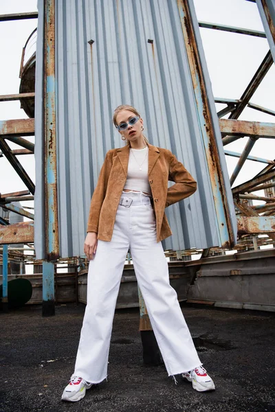 Full length of pretty young model in blue sunglasses posing with hand on hip near rusty construction on rooftop — Stock Photo