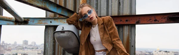 Trendy model in blue sunglasses and stylish outfit posing on rusty rooftop, banner — Stock Photo