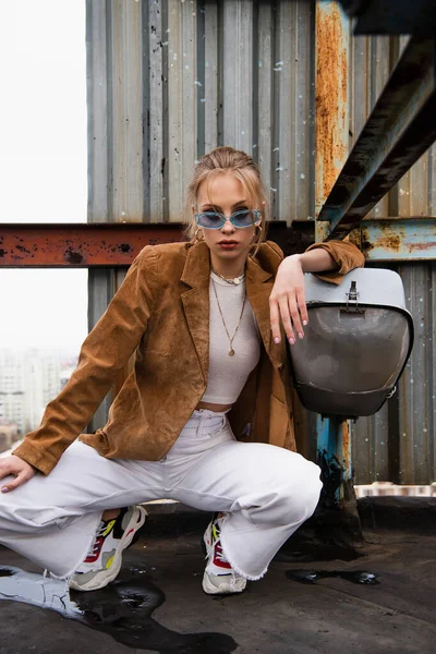 Mujer rubia en gafas de sol azules y ropa de moda sentada mientras posa en la azotea oxidada - foto de stock