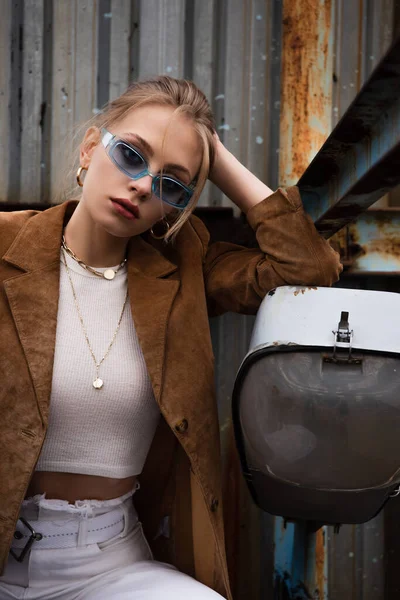 Mujer rubia en gafas de sol azules y chaqueta de gamuza sentada mientras posa en la azotea oxidada - foto de stock