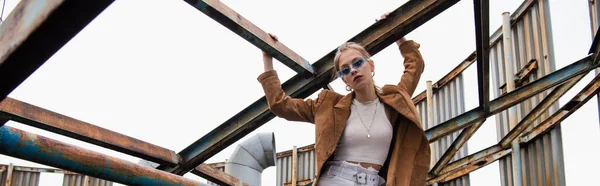 Donna bionda in occhiali da sole blu e giacca scamosciata in posa vicino alla costruzione arrugginita sul tetto, banner — Foto stock