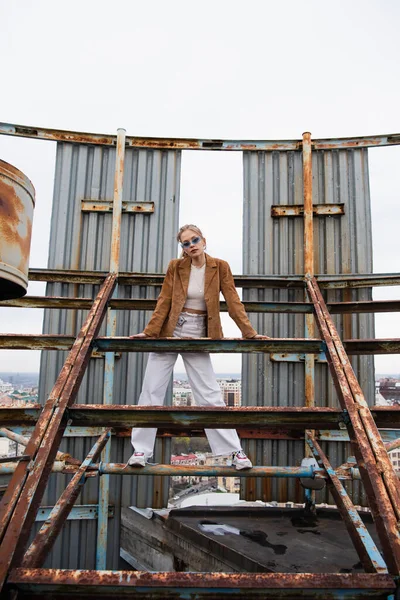 Longitud completa de la mujer joven en gafas de sol azules y traje de moda de pie en la construcción oxidada de la azotea - foto de stock