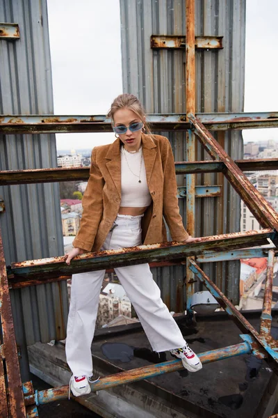 Full length of young woman in blue sunglasses and trendy outfit standing on weathered construction of rooftop — Stock Photo