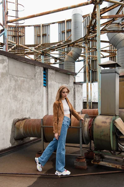 Comprimento total de jovem modelo loira em jeans jeans jeans e tênis elegantes posando no telhado — Fotografia de Stock