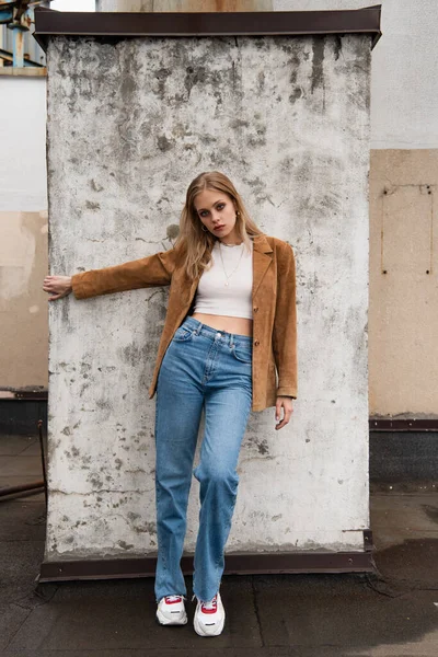 Полная длина блондинка в джинсах джинса, замшевый блейзер и модные кроссовки позируют возле бетонной стены на крыше — стоковое фото