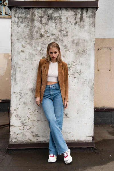 Comprimento total do modelo em jeans, blazer de camurça e tênis elegantes posando com pernas cruzadas perto da parede de concreto no telhado — Fotografia de Stock