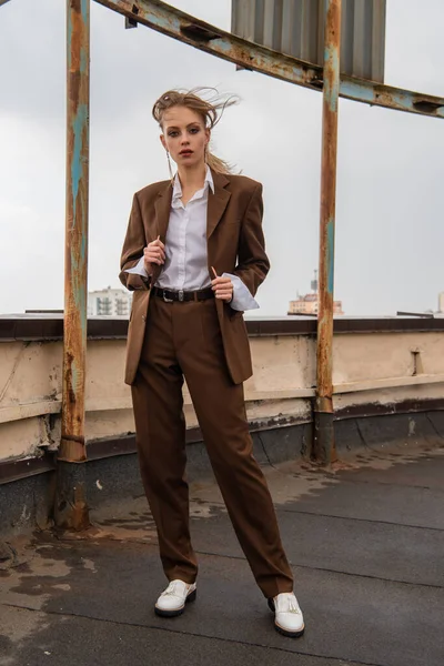Full length of young model in earrings, white shirt and brown suit looking at camera while posing on rooftop — Stock Photo