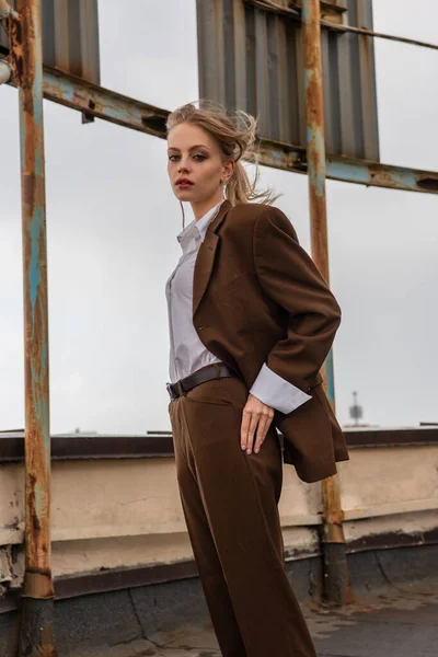 Junges Model in Ohrringen, weißem Hemd und braunem Anzug schaut in die Kamera, während sie auf dem Dach posiert — Stockfoto